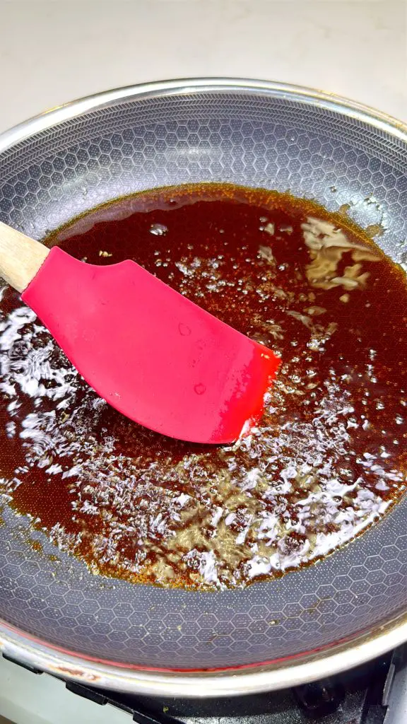 A frying pan on a stove with bubbling brown sauce. A red spatula is stirring the sauce, which appears to be caramelizing. The pan has a textured, non-stick surface.
