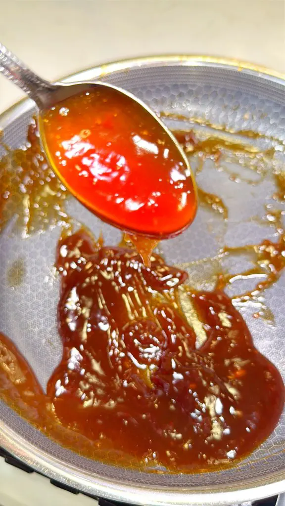 A spoon with red, glossy sauce above a pan containing thick, dark brown sauce. The texture appears sticky and smooth, with reflections of light on the surface.