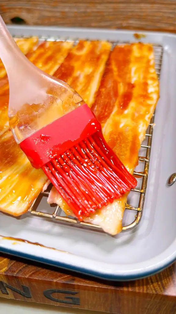 A close-up of bacon slices on a wire rack over a tray. A red silicone brush is being used to apply a shiny glaze over the bacon. The glaze has a rich, glossy appearance.