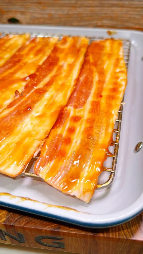 Strips of uncooked bacon coated in a glossy, reddish sauce are placed on a metal rack. The rack is positioned over a white tray, which collects any excess sauce. The bacons marbling is visible through the glaze.