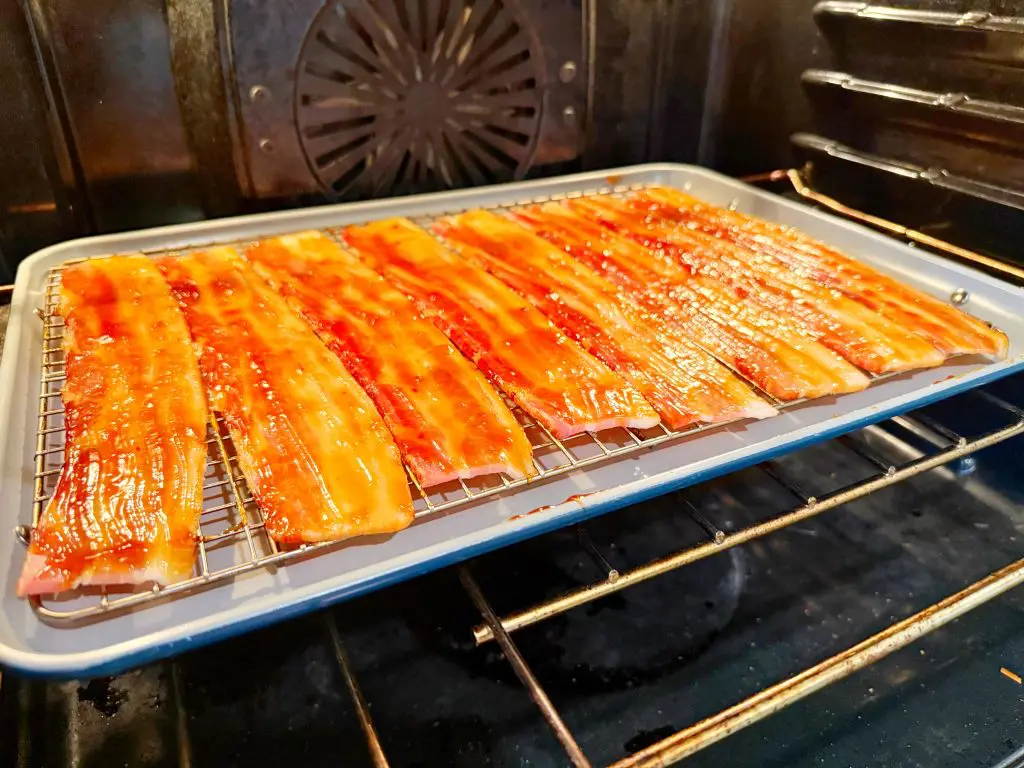 Strips of candied bacon on a wire rack over a baking sheet in an oven. The bacon has a glossy, caramelized appearance.