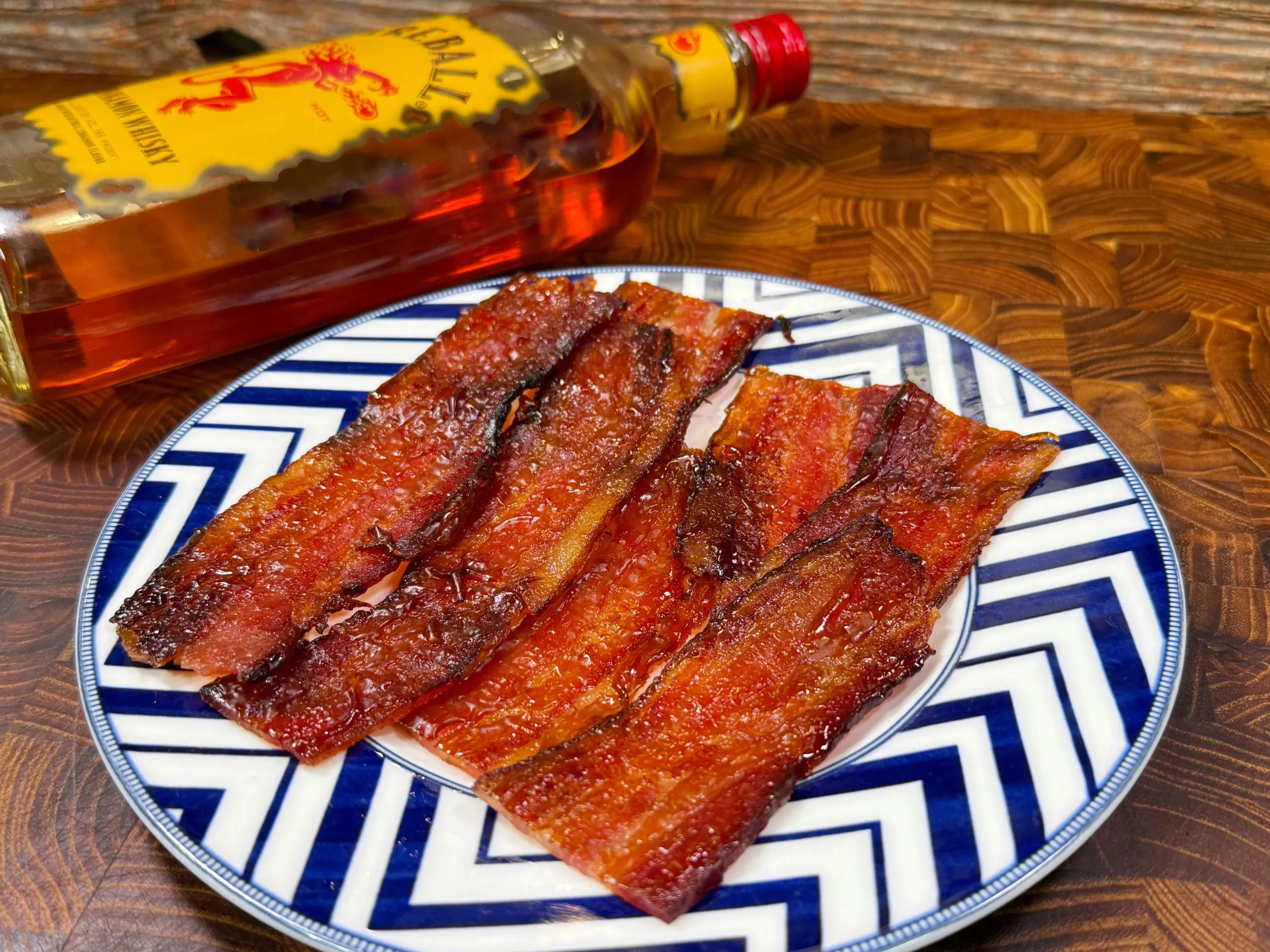 A plate with crispy bacon strips is on a blue and white zigzag patterned dish. A bottle of cinnamon-flavored whiskey lies on its side in the background, on a wooden surface.