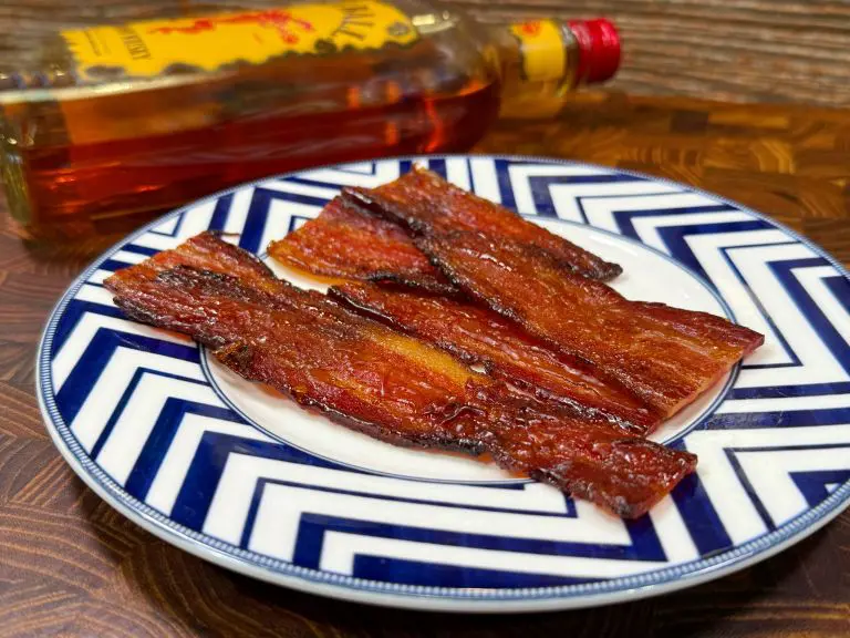 Plate of glazed bacon strips on a blue and white zigzag-patterned plate, with a bottle of Fireball whiskey in the background, on a wooden surface.