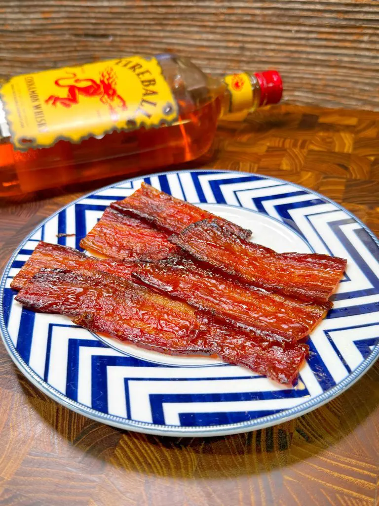 Plate of glazed bacon strips on a blue and white patterned dish. A bottle of Fireball cinnamon whisky lies on its side in the background on a wooden surface for this candied bacon recipe.