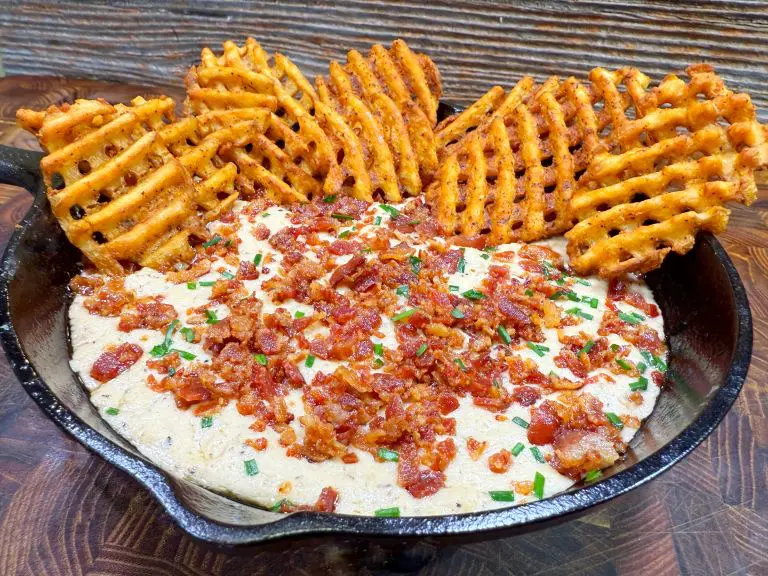 A skillet filled with creamy dip, topped with crumbled bacon and chopped chives. The dish is surrounded by crispy waffle fries standing upright. The background features a rustic wooden wall.