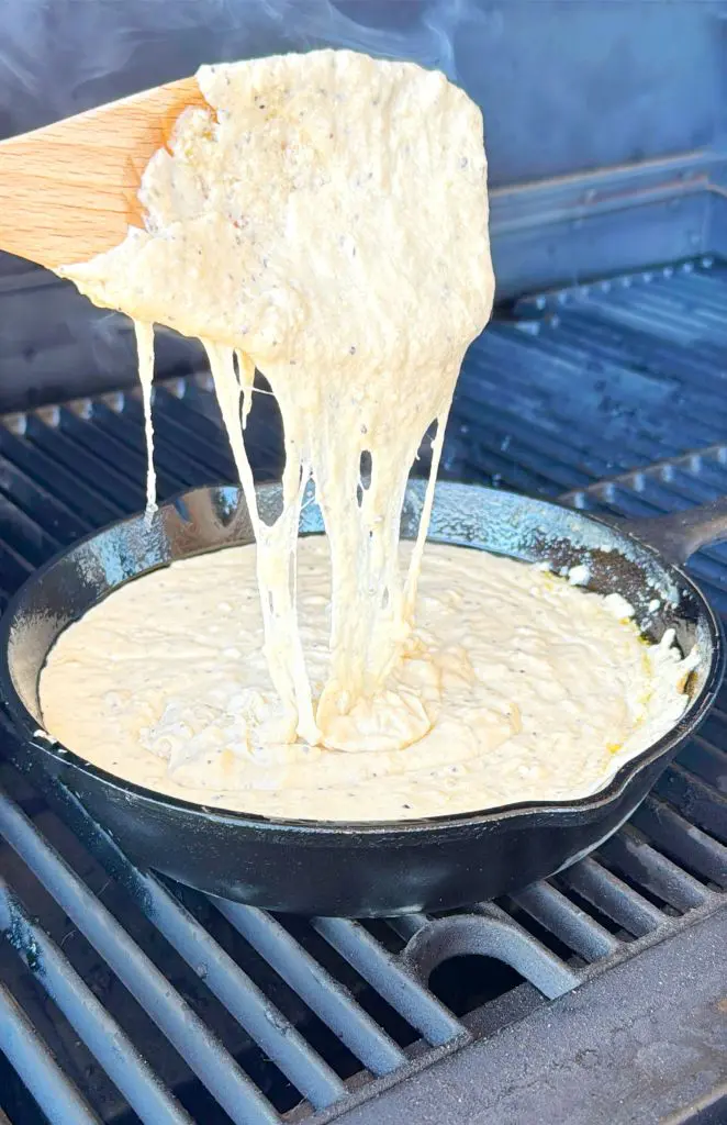 A wooden spatula lifts gooey, melting cheese from a bubbling dip in a cast iron skillet on a grill. The melted cheese stretches, showing its stringy texture.