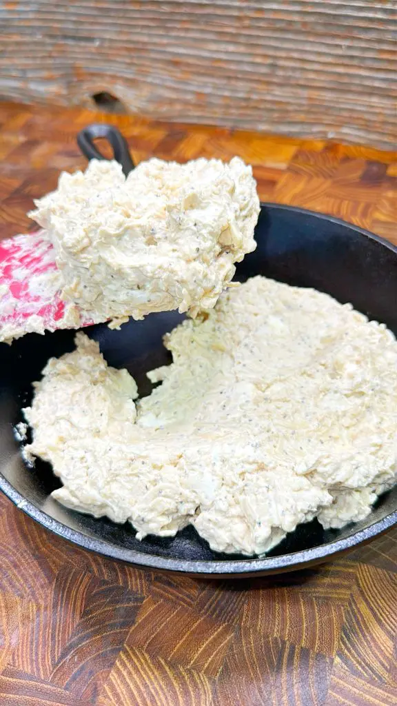 A pan is filled with a creamy, textured mixture being scooped with a red and white spatula. The contents appear thick and light in color, possibly a spread or batter, set against a wooden background. Poutine dip is almost completed