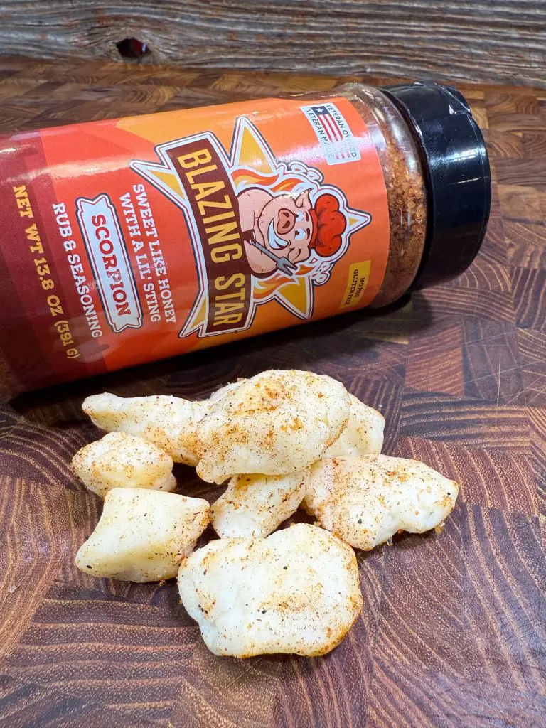 A container of Blazing Star seasoning labeled Scorpion lies on its side, spilling some seasoning powder onto puffed pork skins on a wooden surface. The pork skins are dusted with the seasoning.