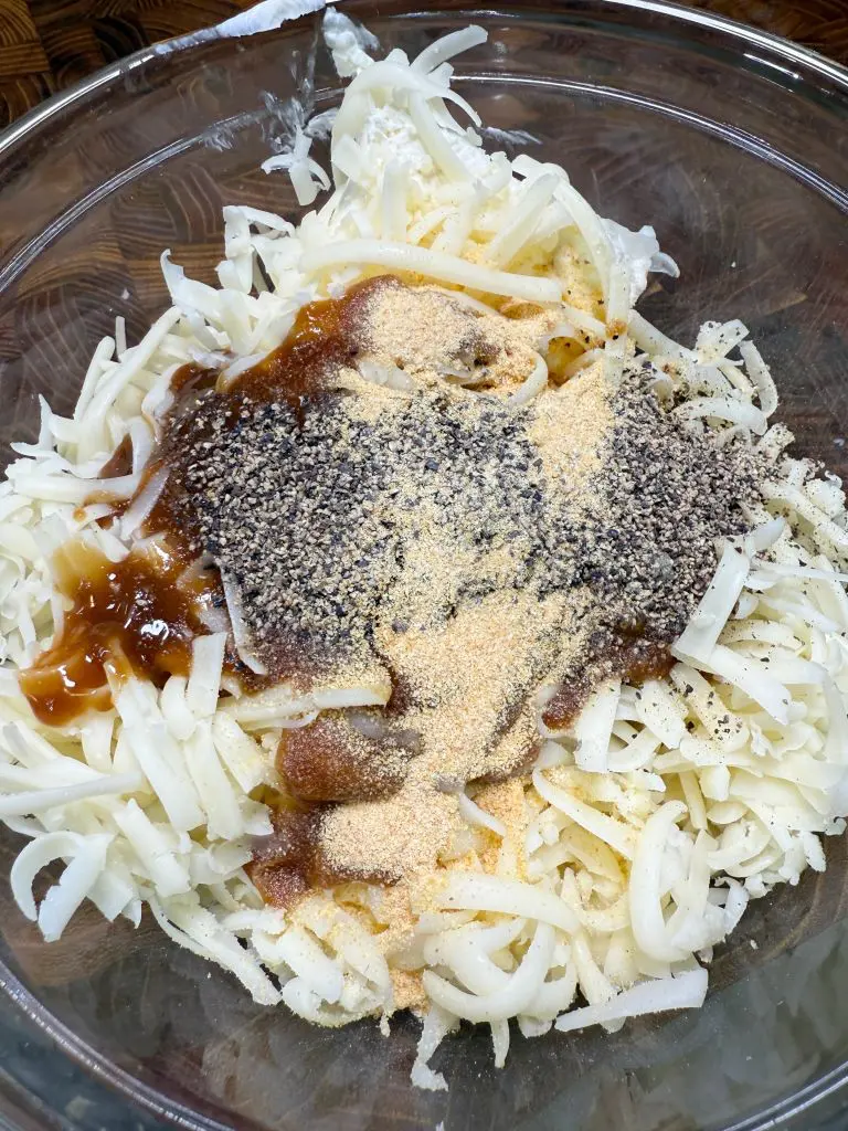 A glass bowl filled with shredded potatoes, grated cheese, black pepper, garlic powder, and a brown sauce, ready to be mixed.
