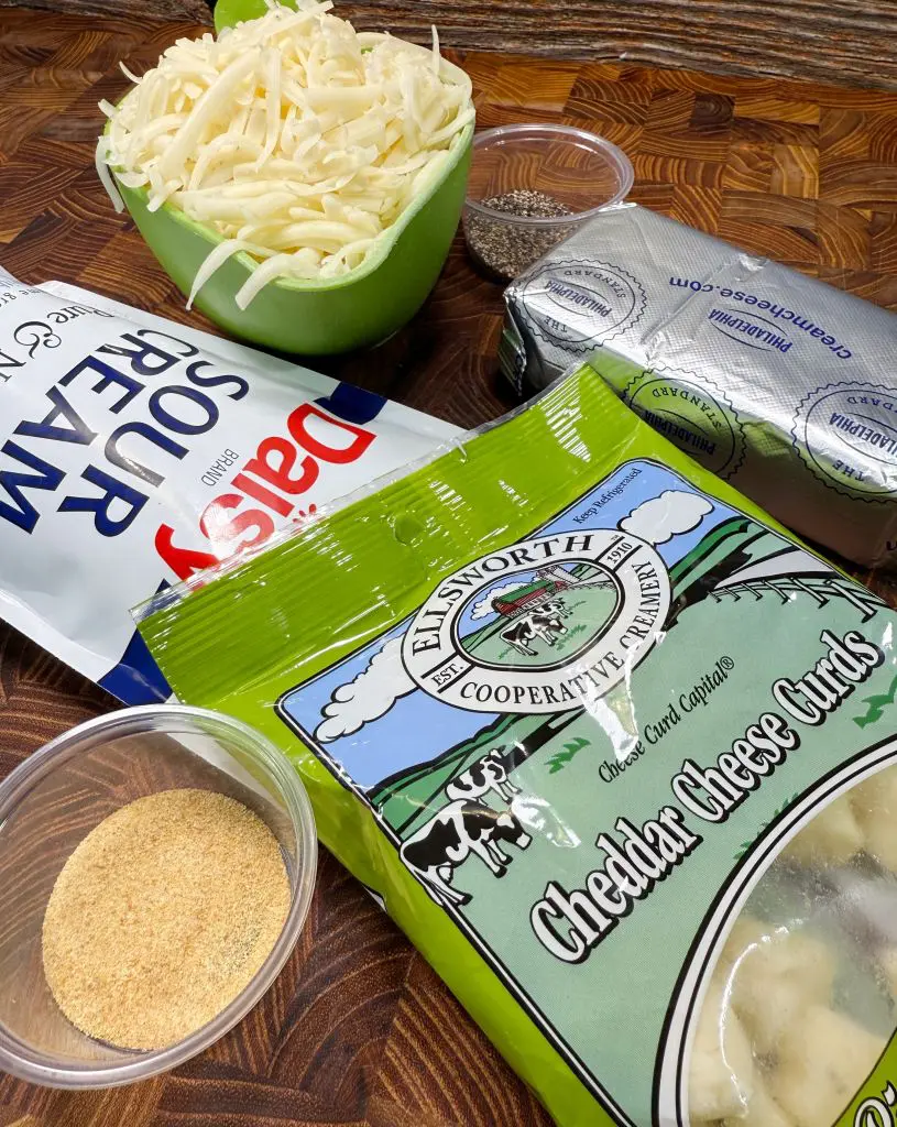 A display of cooking ingredients: a green bowl of shredded cheese, a packet of sour cream, a container of poppy seeds, a packet of cheddar cheese curds, a foil-wrapped package of cream cheese, and a small cup of garlic powder on a wooden surface.