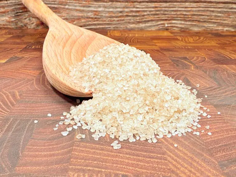 A wooden spoon lies on a wooden surface, spilling a heap of coarse sea salt crystals. The background is a textured wood pattern, enhancing the rustic appearance.