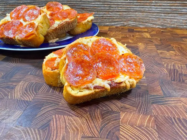 Slices of bread topped with melted cheese and pepperoni are served on a wooden surface. A plate with additional slices is partially visible in the background. The setup has a rustic, textured backdrop.