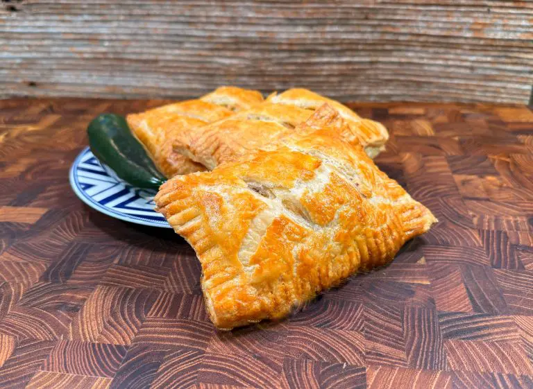 A plate with three golden-brown pastries, puffed and baked, is set on a wooden surface. A whole green jalapeño pepper lies beside the pastries, adding a touch of color. The plate features a blue and white geometric pattern.