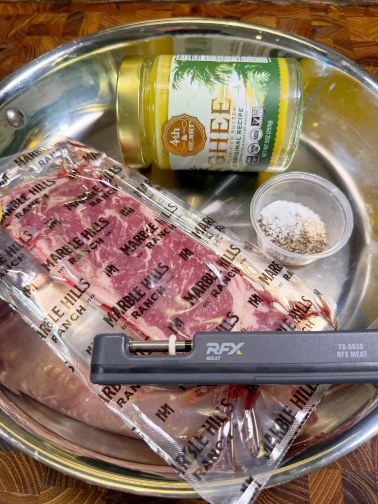 A stainless steel pan holds a vacuum-sealed package of raw meat labeled Marble Hills Ranch, a jar of Fourth & Heart ghee with a vanilla flavor, a small container of salt and pepper, and a meat thermometer.