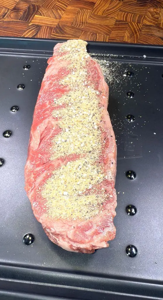 Raw beef steak seasoned with a mixture of spices, displayed on a black metal tray with a textured wooden background. The seasoning is spread over the top surface of the steak.