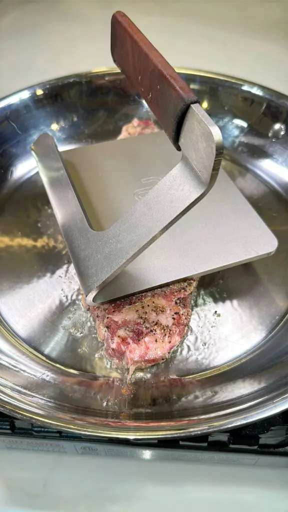 A stainless steel burger press with a wooden handle is pressing a seasoned beef patty on a frying pan. The patty is sizzling, with visible juices and pepper.