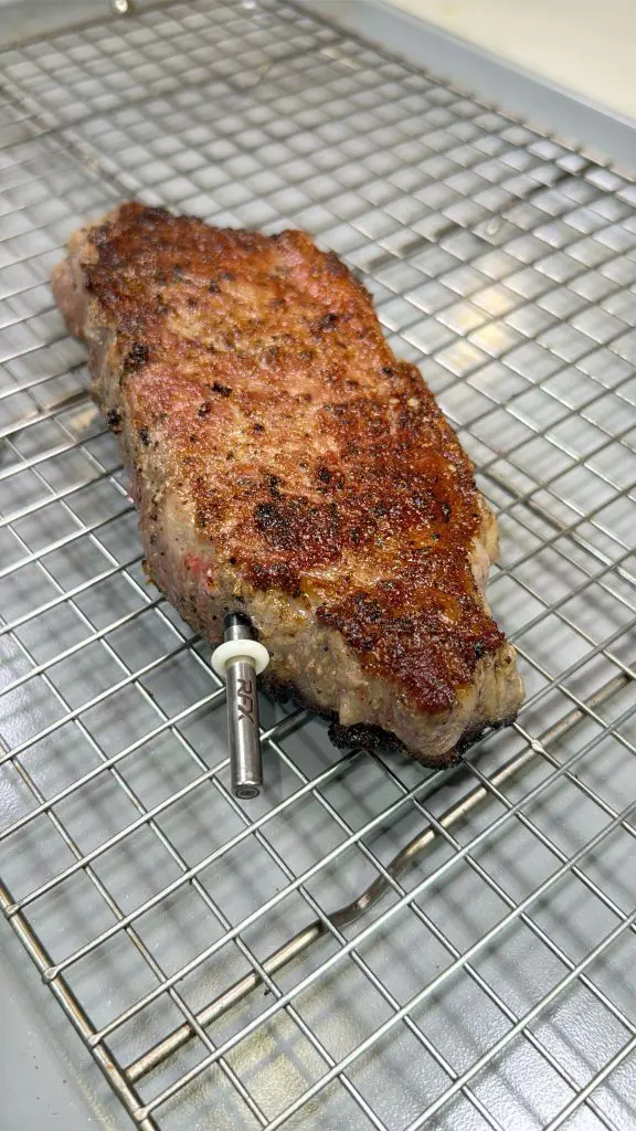 Grilled steak resting on a wire rack with a meat thermometer inserted into its side. The steak has a seared, textured surface and is placed on a metal grid over a kitchen countertop.
