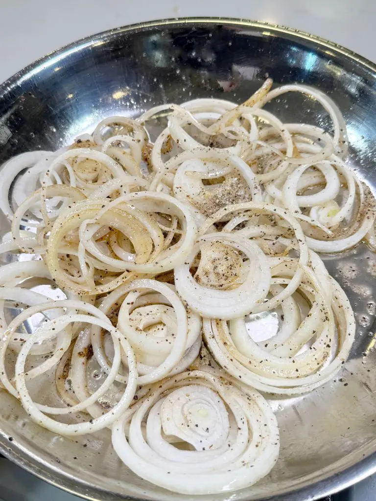 Sliced onions cooking in a stainless steel pan, seasoned with cracked black pepper. The onions are slightly translucent, suggesting they are being sautéed. The pan is on a stove with a reflective surface.