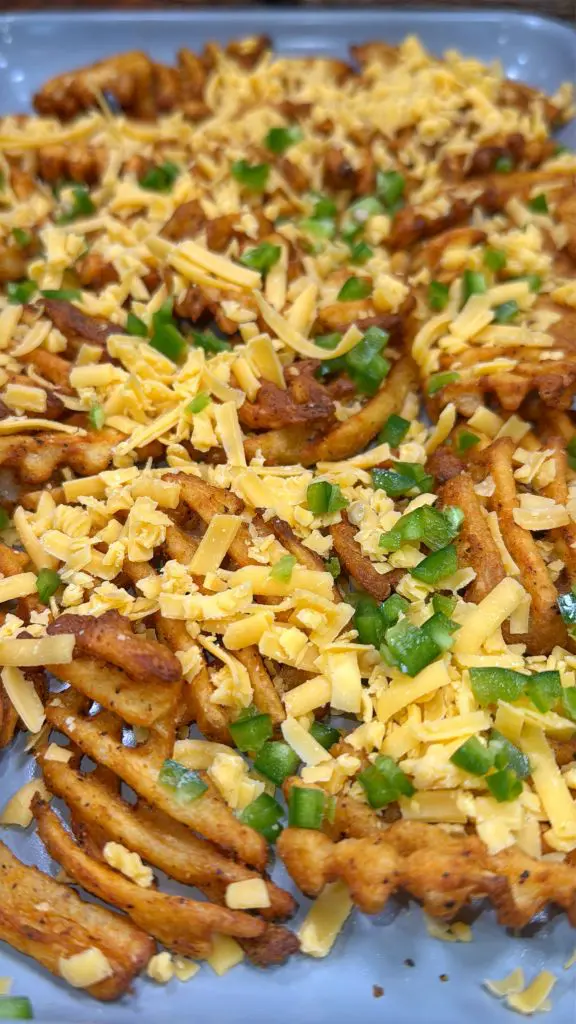 A tray of loaded french fries / waffle fries topped with shredded cheese and chopped green peppers. The golden-brown fries are seasoned and covered with melted cheese, creating a savory and colorful dish.