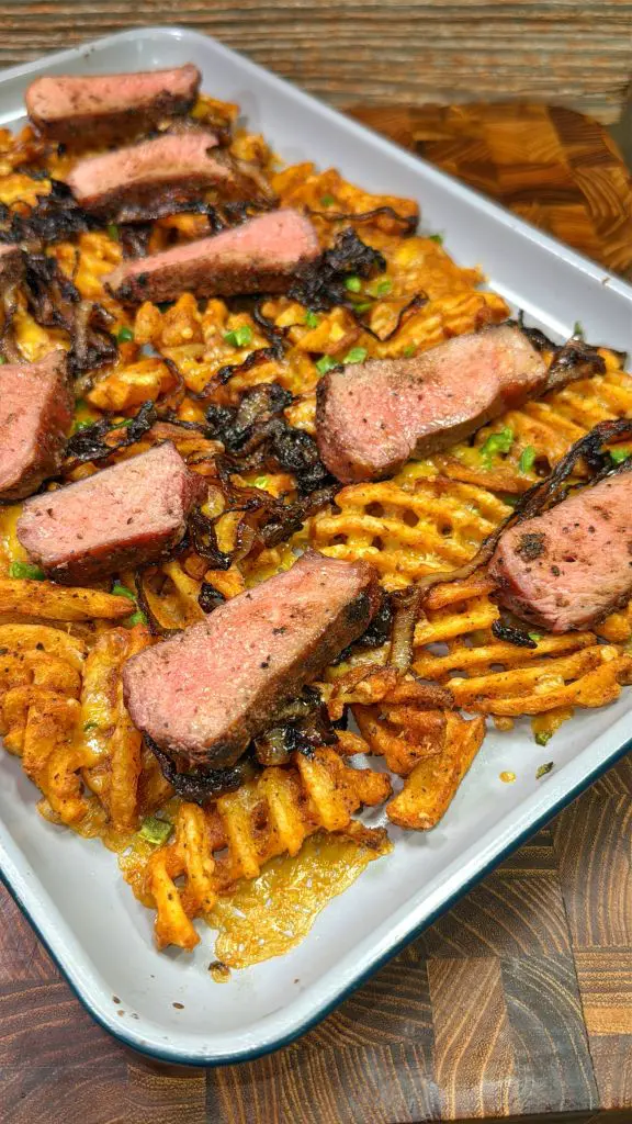 A tray of waffle fries topped with sliced steak, garnished with caramelized onions and chopped parsley. The steak is cooked to medium doneness, and the fries are golden and crispy. The background shows a wooden surface.