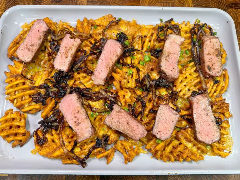 A white tray holds waffle-cut fries topped with slices of medium-cooked steak and caramelized onions. The fries are slightly crispy and sprinkled with chopped herbs.