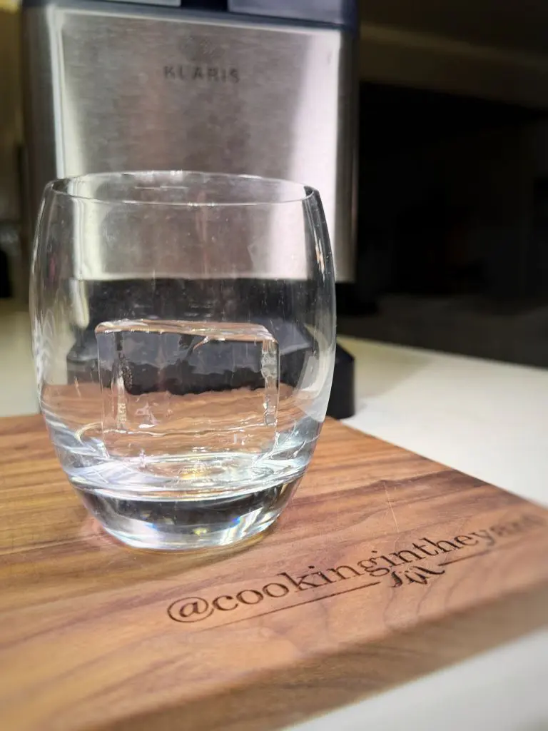 A clear glass with a large square ice cube inside rests on a wooden board. The board is engraved with @cookingintheyard. A metallic appliance is blurred in the background.