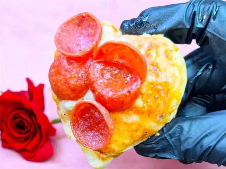 A gloved hand holds a heart-shaped mini pizza topped with pepperoni slices. In the background, theres a red rose on a pink surface.