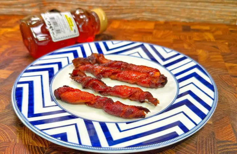 A plate with three strips of glazed, crispy bacon on a blue and white zigzag patterned plate. A small bottle of honey lies on its side in the background on a wooden surface.