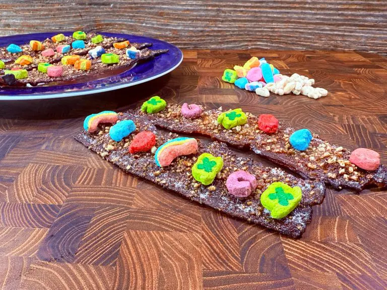 Chocolate bacon strips topped with colorful cereal marshmallows are displayed on a wooden surface. A blue plate with similar strips is in the background, alongside a small pile of marshmallows. The surface features a rich wood grain pattern.