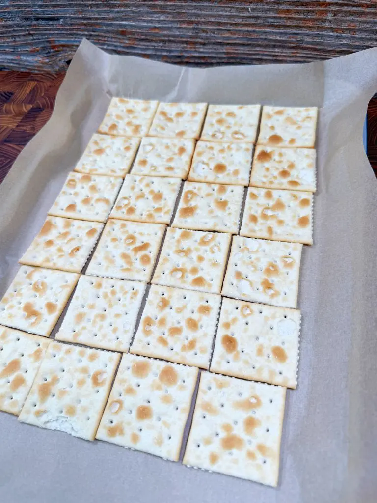 A grid of square saltine crackers arranged neatly on a sheet of parchment paper. The crackers have a light, baked texture with small holes and brown spots, set against a rustic wooden background.