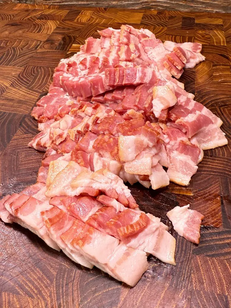 Chopped pieces of raw bacon are spread out on a wooden cutting board with a rich grain pattern. The bacon is pink with visible layers of fat, and the board shows a textured surface.