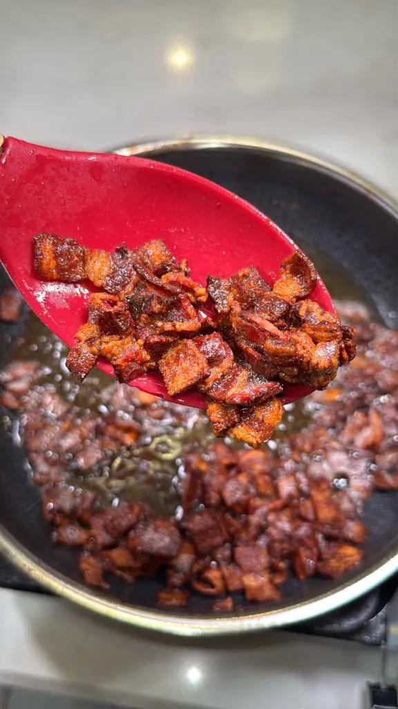 Close-up of crispy, browned bacon pieces on a red spatula held over a pan. The pan is filled with more cooked bacon pieces glistening in oil. The background is a light-colored stovetop surface.