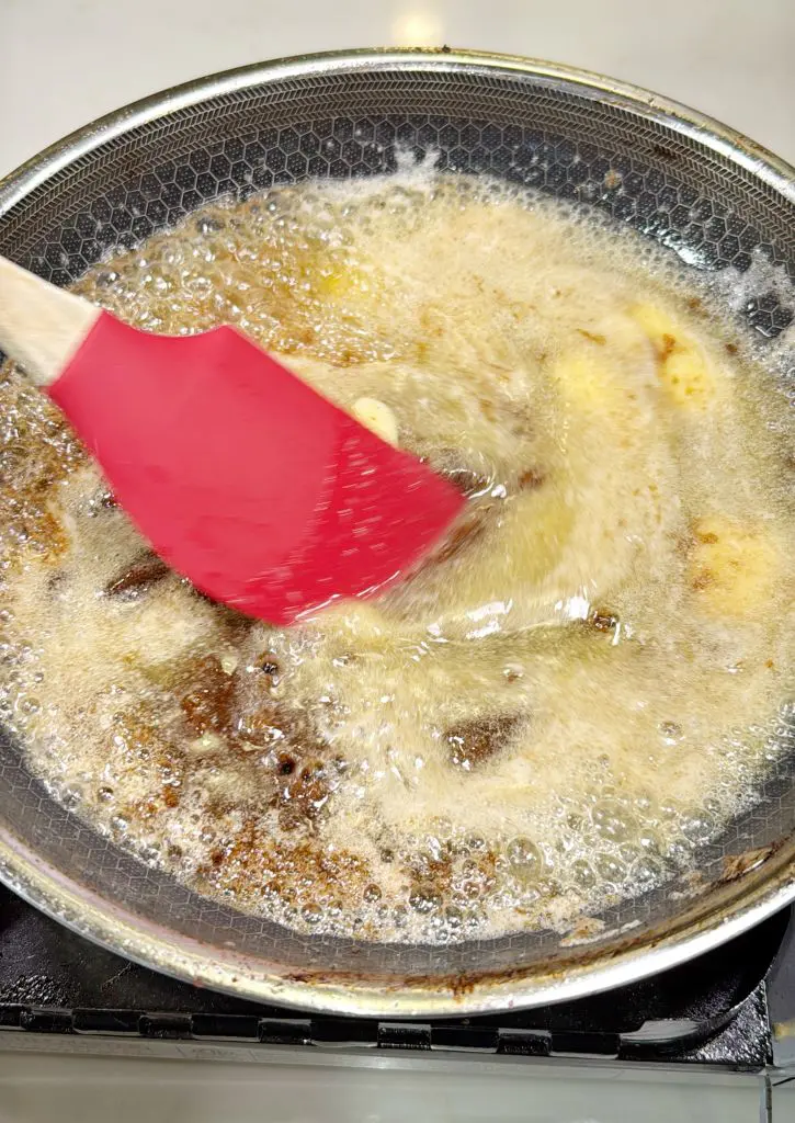 A red spatula stirs crispy bacon bits in a hot pan of bubbling oil and foam on a stove. The motion creates a ripple effect across the surface of the liquid. The pan has a non-stick, textured bottom.