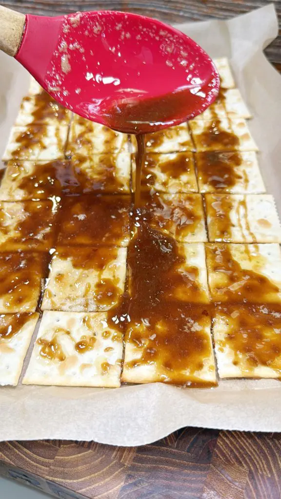 Red spoon drizzling caramel sauce over a layer of saltine crackers on parchment paper, set on a wooden surface.