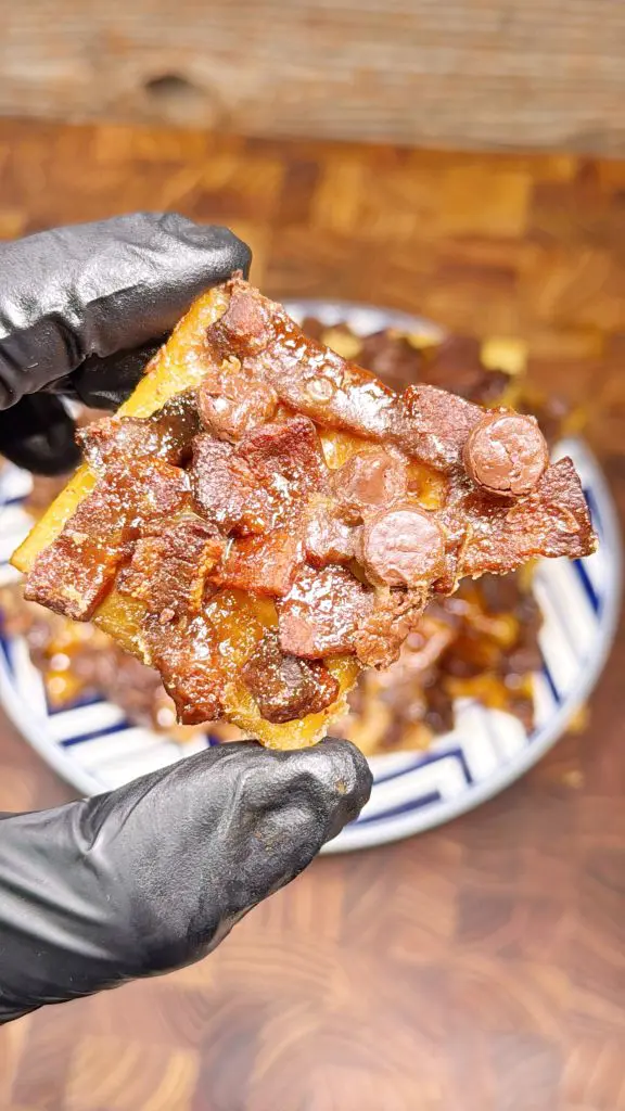 A gloved hand holding a savory square treat topped with chunks of bacon and small round pieces, possibly chocolate chips, over a blurred plate of similar treats on a wooden surface. This recipe is for bacon crack