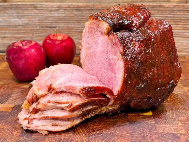 Close-up of a sliced, glazed ham on a wooden cutting board, paired with two red apples in the background. The ham has a shiny, caramelized exterior and a pink, tender interior, suggesting a savory, flavorful dish.