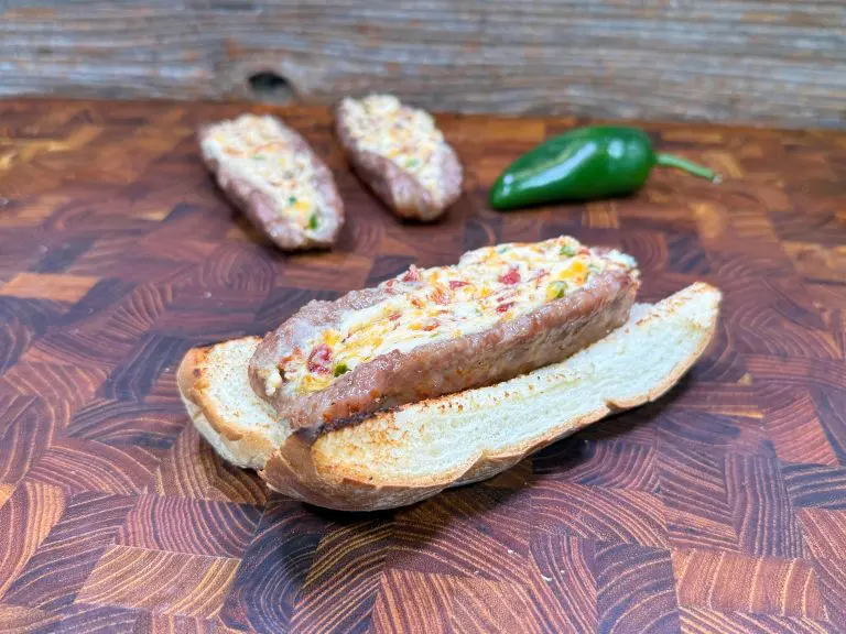 A grilled sandwich sits open-faced on a wooden surface. Its filled with melted cheese, diced jalapeños, and crumbled bacon. Two more sandwiches and a whole jalapeño are slightly blurred in the background.