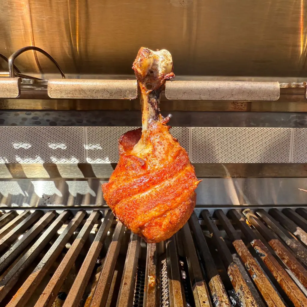 A cooked turkey leg, seasoned and browned, is upright on a grill. The grills metal bars and the ovens interior are visible in the background. This image is of a bacon wrapped chicken drumstick