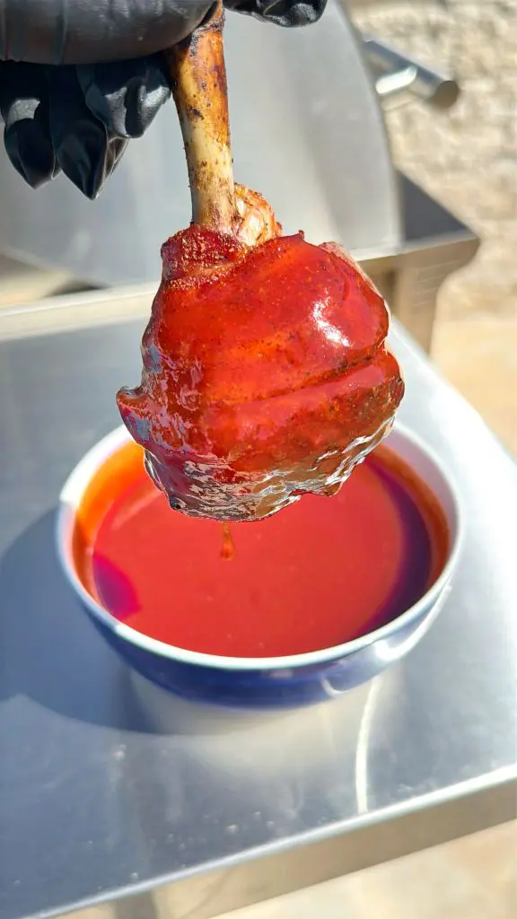 A gloved hand holds a piece of meat coated in shiny, red barbecue sauce above a bowl filled with the same sauce. The background features a stainless steel surface, hinting at an outdoor cooking area. Delicious bacon wrapped chicken drumsticks