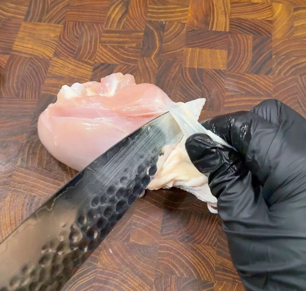 A person wearing black gloves is holding a knife, skinning a piece of raw chicken on a wooden cutting board. The blade is angled to remove the skin neatly from the meat.