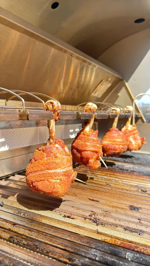 Grilled chicken drumsticks wrapped in bacon, suspended on hooks inside a barbecue grill. The grill grates below are slightly charred, and the chicken has a golden-brown, crispy appearance. Sunlight brightens the scene.