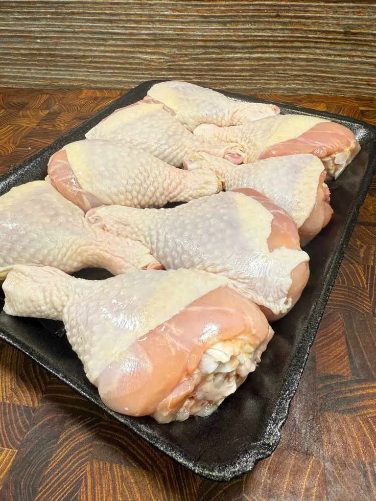 Tray of raw chicken drumsticks and thighs with pale skin, arranged neatly on a dark platter. The wooden surface beneath has a warm, textured appearance.