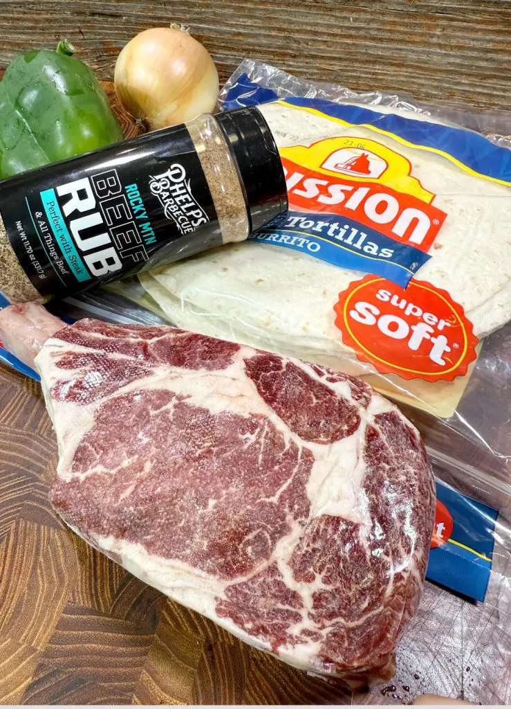 A marbled raw ribeye steak, a pack of Mission tortillas, a bottle of beef rub seasoning, a green bell pepper, and a yellow onion on a wooden surface—everything you need to create the ultimate Cheesesteak Quesadilla.