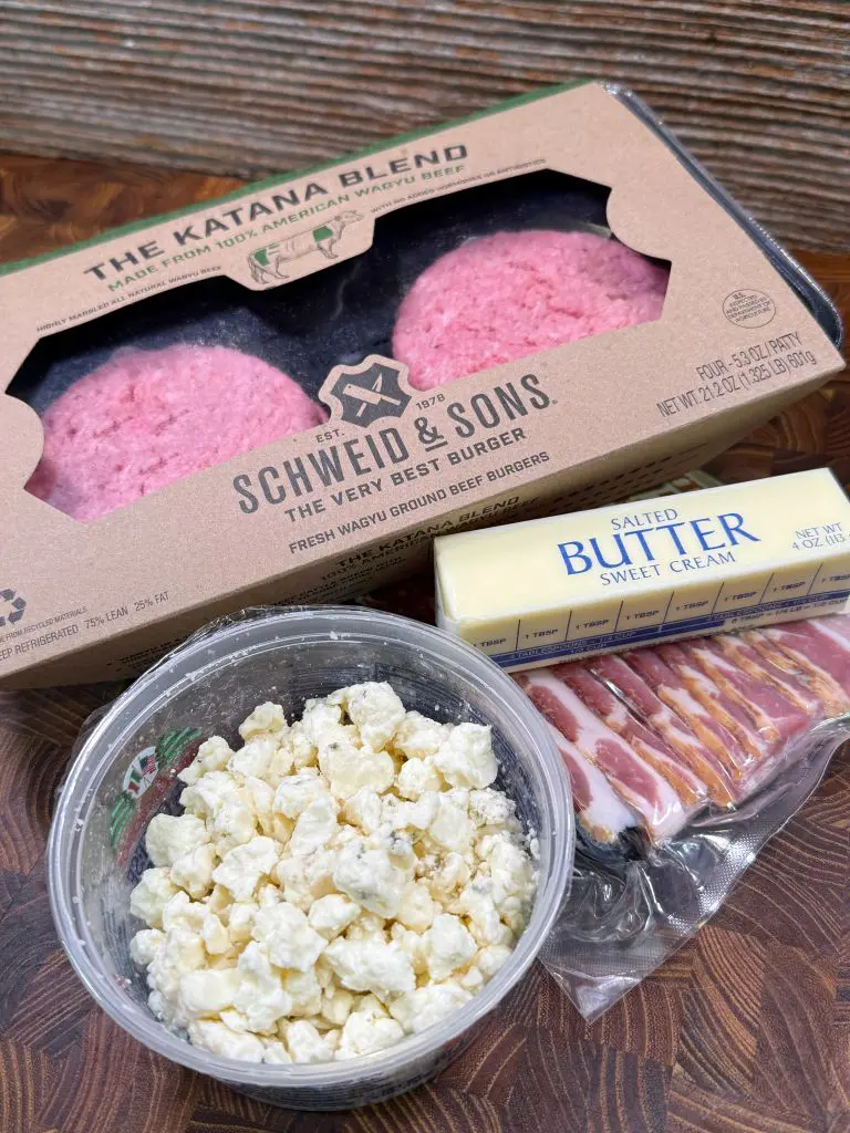 A package of Schweid & Sons ground beef patties, a stick of salted sweet cream butter, a pack of sliced bacon, and a container of crumbled cheese on a wooden surface.