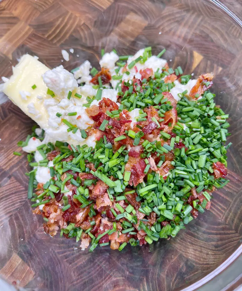 A glass bowl containing chopped chives, crumbled bacon, cubes of butter, and cream cheese on a wooden surface, ready to be mixed.