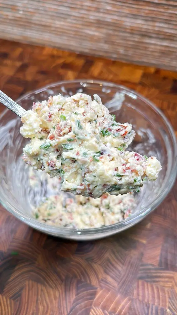 A spoon lifts a mixture of finely chopped herbs, bacon bits, and a creamy base from a glass bowl, set on a wooden surface.