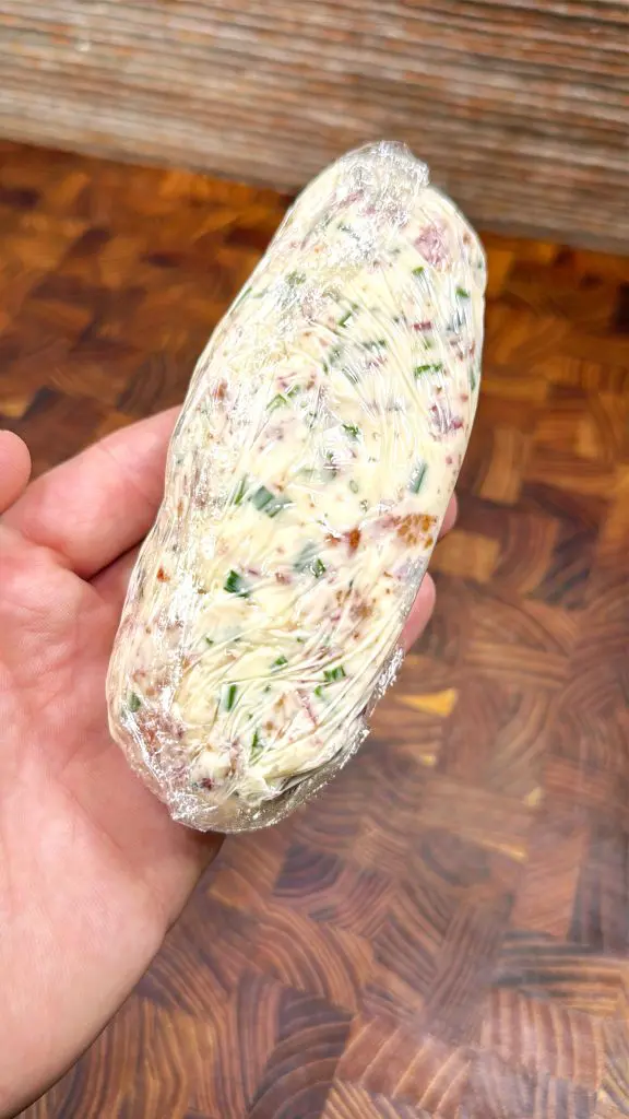 A hand holds a log of compound butter mixed with herbs and possibly bits of dried meat, tightly wrapped in plastic wrap, over a patterned wooden surface.