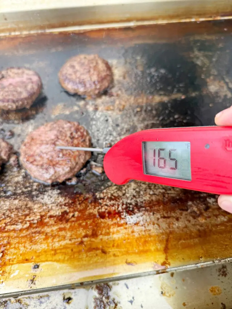 A hand holds a red digital meat thermometer inserted into a hamburger patty on a grill. The thermometer displays a temperature of 165°F. Several other patties are also cooking on the greasy grill surface.