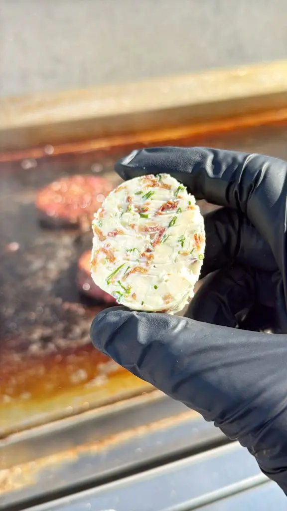 A gloved hand holds a round slice of compound butter with visible herbs and bits of bacon; a grill with burgers cooking is blurred in the background.