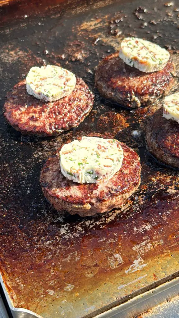 Four beef patties are cooking on a grill, each topped with a round slice of herb butter. The surface of the grill is well-used and browned from the heat.